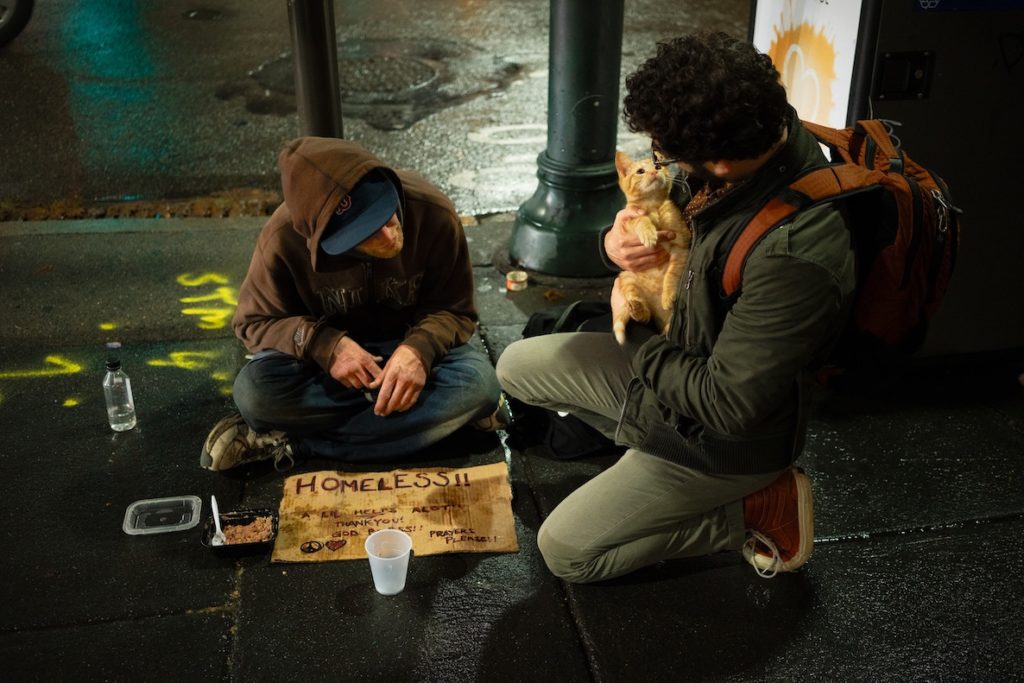 A man stops to play with a homeless man's cat on a rainy night in the city.