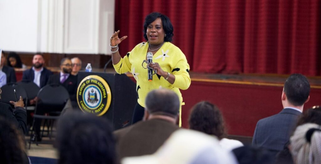 Mayor of Philadelphia Cherelle Parker, a Black woman with black hair, stands with a microphone and her large frame black and white glasses in one hand. She's in front of an auditorium crowd wearing a yellow sweater with white flowers. Her arm is raised and she is mid-speech.